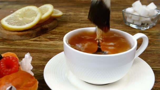 Bag of tea brewed in white ceramic mug, dipped in boiled hot water and two pieces of refined sugar added and stir with teaspoon. Sliced lemon and cake lies on wooden table. Close-up. — Vídeo de stock