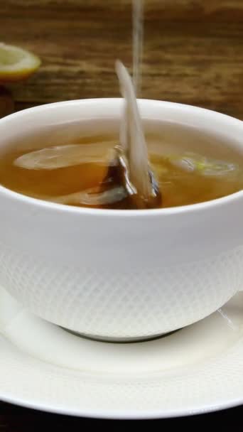 En påse te bryggt i vit keramisk mugg, doppat i kokt varmt vatten och två bitar raffinerat socker tillsatt. Skivad citron ligger på träbordet. Vertikalt skott. — Stockvideo