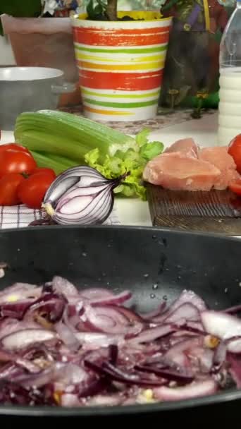 Mesa en cocina doméstica. Preparación de alimentos mediterráneos. Sartén con cebolla en rodajas. Chef cortando tomate para hacer sabrosa salsa con carne de pollo, cebolla, apio. Vídeo vertical. — Vídeos de Stock