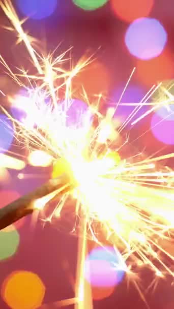 Brilhante sparkler queima contra o pano de fundo de guirlanda piscando multicolorido desfocado. Cenário desfocado. Feliz Natal ou aniversário conceito de férias. Vídeo vertical. — Vídeo de Stock