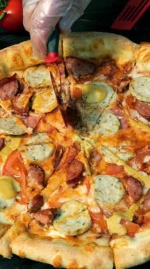 Elleri kesilmiş pizza, dilimler halinde, sosis, domates ve peynirle, özel bıçakla, kesici tekerlekle. Tahta masada domates ve rendelenmiş peynir de var. Dikey biçim.