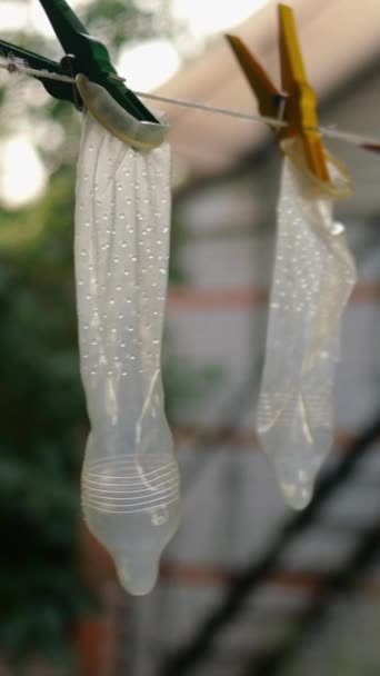 Transparante latex condooms hangen op waslijn in de achtertuin van het huis, bevestigd met wasknijpers en schommel in lichte bries. Misschien zijn ze gewassen om weer gebruikt te worden. Verticaal formaat. Buiten. — Stockvideo