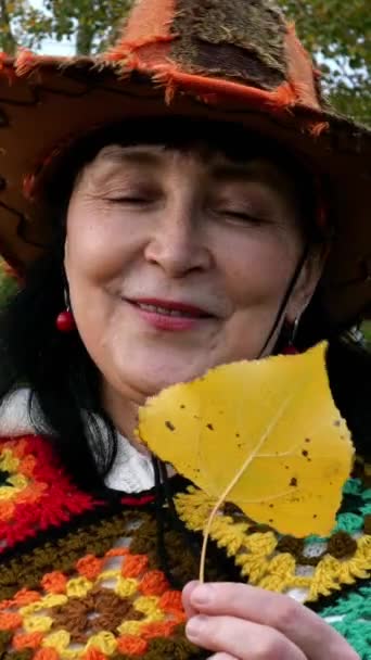 Elegante volwassen vrouw in mooie heldere kleren praat emotioneel in het najaarspark, houdt geel blad vast en glimlacht. Verticale video. Close-up. — Stockvideo