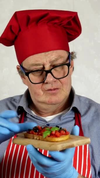 Chef adulte majeur d'origine caucasienne au chapeau de chef rouge, dégustation de bruschetta aux tomates et au basilic, dans la cuisine domestique. Vidéo verticale. Gros plan. — Video