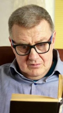 Yaşlı yetişkin bir adam kafkas kökenli, odasındaki deri koltukta oturuyor, kitap okuyor ve kırmızı bardaktan çay ya da kahve içiyor..