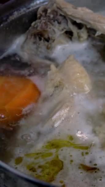 Cocinar en agua hirviendo el caldo de pollo graso de carne y huesos con cebollas y zanahorias para la preparación de la sopa, en una cacerola. Cocina doméstica. Vídeo vertical. Primer plano. — Vídeo de stock