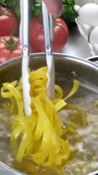 シェフは鍋でパスタを調理.家庭用キッチンでイタリアのタリアテッレを準備します。鍋にパスタを混合トングで調理します。キノコ、卵、トマト。垂直ビデオ. — ストック動画
