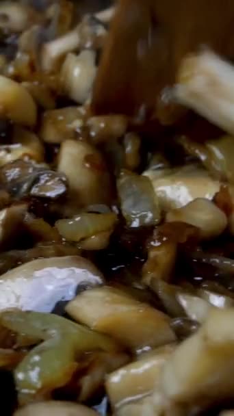 Finely chopped mushrooms are fried in pan in boiling vegetable or olive oil and mixed thoroughly with kitchen spatula. Domestic kitchen. Dolly shot. Vertical video. Close-up. — Stock Video