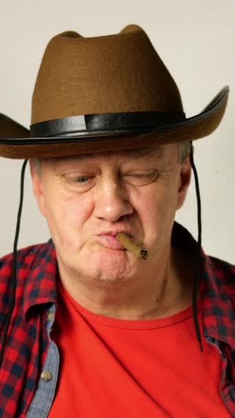 Hombre mayor de etnia caucásica en un sombrero de vaquero con una mirada descarada, fuma un cigarro y sopla humo hacia la cámara. — Vídeo de stock