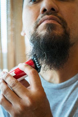 Asyalı adam elektrikli tıraş makinesiyle sakal tıraş ediyor..