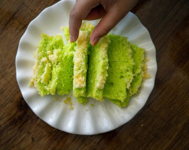 Beyaz tabakta tereyağı kırıntıları olan pandan yeşili kek. Parmakları da bir parça alıyor. Üst görünüm.