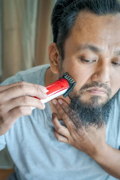 Asian man shaving beard with electric razor machine. High angle view.