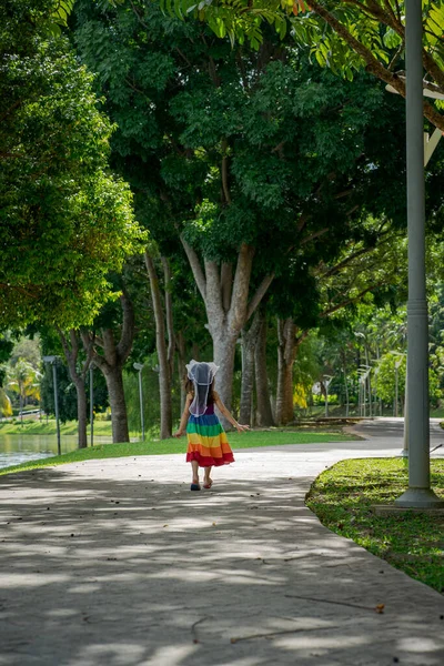 A little girl in a rainbow dress fun running on the concrete path in the park. View from behind.