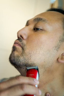 Asyalı adam elektrikli tıraş makinesiyle sakal tıraş ediyor..