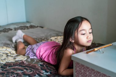 Küçük sevimli kız yatakta oturmuş cep telefonuyla çizgi film izliyor.