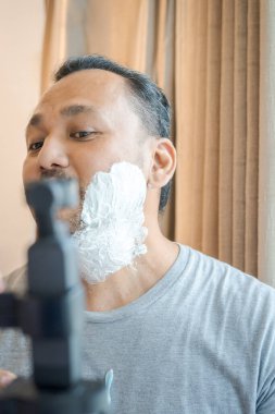 Asyalı bir adamın yüzüne tıraş kremi sürüyorum. Adam kamerada tıraş işlemini kaydediyor..