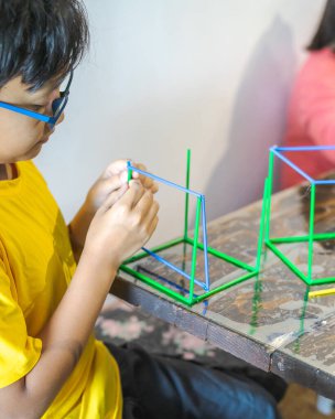 Genç bir çocuk yapıştırıcı tabancasıyla renkli plastik çubuklar yapıyor. Geometrik figürler oluşturmak ve evde matematik öğrenmek eğlenceli.