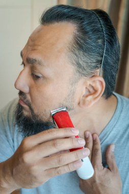 Asyalı adam elektrikli tıraş makinesiyle sakal tıraş ediyor..