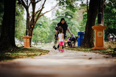 Yürümeye yeni başlamış anne tesettüre binmiş, arka planda bebek arabası tutuyor..
