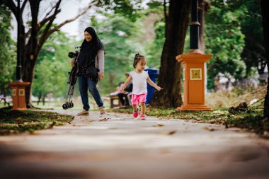 Yürümeye yeni başlamış anne tesettüre binmiş, arka planda bebek arabası tutuyor..