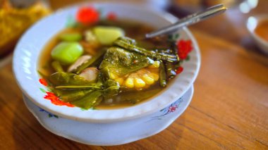 Yapraklı yeşillikler, mısır ve patates parçaları içeren Sayur asem adlı karışık sebze tamarind çorbası. Sıcak, ahşap bir masaya kurulmuş sağlıklı bir yemek, sıcaklığı ve birlikteliği davet ediyor.