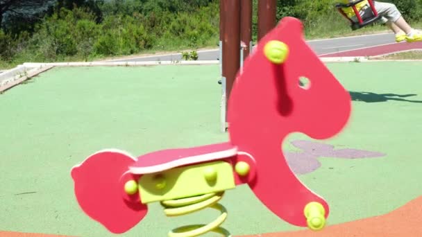 Toy Horse, Un enfant se balançant sur la balançoire derrière un cheval jouet se balançant dans une aire de jeux — Video