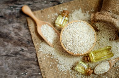 Kuru organik beyaz susam tohumlarının üst görüntüsü ahşap kasede bulanık arka planda bazıları çuval kumaş ve grunge zemininde cam şişede susam yağı ve koyu kahverengi tonda.