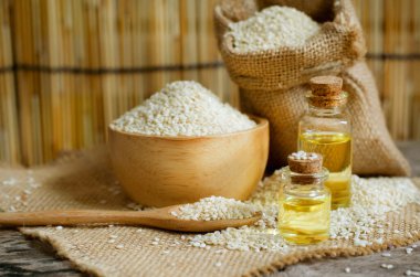 Closed up organic white sesame seed oil in glass bottle and some seed in wooden bowl, spoon and sack bag in soft brown tone. Concept of food extract or agricultural product concept