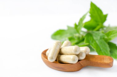 Kapsüle kapsül içinde yeşil Tayland bitkisi tozu, tahta kaşıkta beyaz arka planda bulanık yeşil bitki yaprağı. Doğal ilaçlar ya da kimyasal alerjiden korunmak için alternatif tedavi seçenekleri