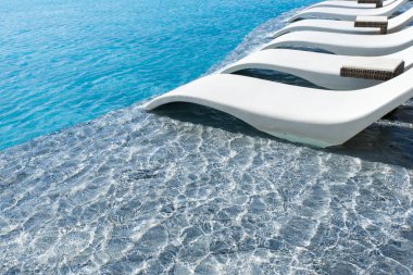 Kenarında beyaz şezlong olan bir havuz. Sandalye havuzun kenarındadır ve suyun içinde konumlandırılmıştır. Havuz mavi bir suyla çevrilidir.