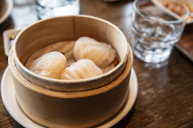 Buharlı köfteler bambu buharında yavaşça dinleniyor. Tahta masa temiz cam ve ince gölgeler barındırır. Yumuşak ışık hassas, şeffaf derileri vurgular. Yemek blogları ve restoran menüleri için mükemmel.