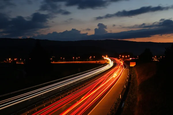 ถนนที่มีการจราจรทางรถยนต์ในเวลากลางคืนพร้อมไฟเบลอในตอนพระอาทิตย์ตก — ภาพถ่ายสต็อก