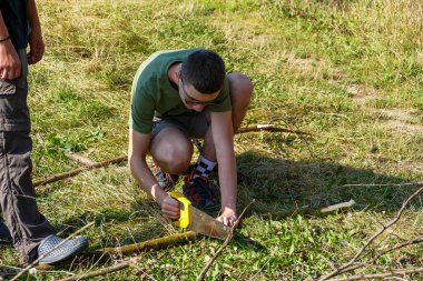 Koyu saçlı adam çömelmiş, ağaç kesiyor, dalları kesiyor, testereyi tutuyor, şenlik ateşi yakıyor, genç, konsantre, şafak, çayır otu, düz, yürüyüş, seyahat, ağaçlar, yaz