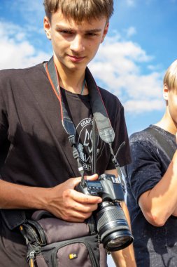 Kahverengi gözlü, kameralı bir gencin portresi, bize bakıyor, gülümsüyor, esmer, yakışıklı, turist, model, açık hava, mavi gökyüzü, bulutlar