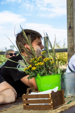 Koyu renk saçlı genç çocuk, tahta çardakta, bukette, kır çiçeklerinde, kurumuş çiçeklerde, doğada, profilde, masada oturuyor.