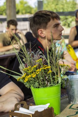 Koyu renk saçlı genç çocuk, tahta çardakta, bukette, kır çiçeklerinde, kurumuş çiçeklerde, doğada, profilde, masada oturuyor.