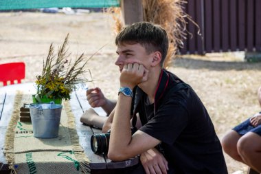 Koyu renk saçlı genç çocuk sokağın tahta çardağında oturuyor, elinde kamera, fotoğrafçı, yanağını destekliyor, profil çıkarıyor, ders dinliyor, bir grup çocuk...