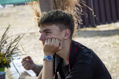 Koyu saçlı genç çocuk ahşap masada oturuyor, yanağı elinde, sokak ahşap çardağı, bir buket kurutulmuş çiçek, sarı cilt yüzeyi, kır çiçekleri, doğa, profil, alektür dinleyerek.