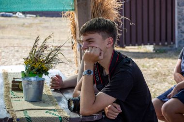 Koyu renk saçlı genç çocuk sokağın tahta çardağında oturuyor, elinde kamera, fotoğrafçı, yanağını destekliyor, profil çıkarıyor, ders dinliyor, bir grup çocuk...