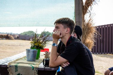 Koyu renk saçlı genç çocuk sokağın tahta çardağında oturuyor, elinde kamera, fotoğrafçı, yanağını destekliyor, profil çıkarıyor, ders dinliyor, bir grup çocuk...