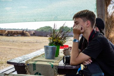 Koyu renk saçlı genç çocuk sokağın tahta çardağında oturuyor, elinde kamera, fotoğrafçı, yanağını destekliyor, profil çıkarıyor, ders dinliyor, bir grup çocuk...