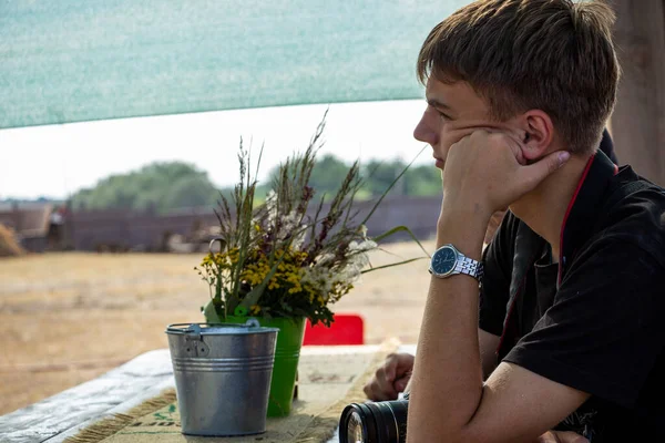 Koyu renk saçlı genç çocuk sokağın tahta çardağında oturuyor, elinde kamera, fotoğrafçı, yanağını destekliyor, profil çıkarıyor, ders dinliyor, bir grup çocuk...