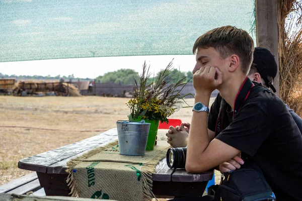 Koyu renk saçlı genç çocuk sokağın tahta çardağında oturuyor, elinde kamera, fotoğrafçı, yanağını destekliyor, profil çıkarıyor, ders dinliyor, bir grup çocuk...