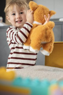 Neşeli küçük bir çocuk evde peluş bir oyuncak köpekle oynuyor. Çocuk gülümsüyor ve yaratıcı oyunlar oynuyor. Duygularını ve merakını ifade ediyor. Kapalı alan atmosferi. 
