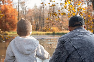 Bir sonbahar parkında sakin bir gölün kenarında duran bir adam ve küçük çocuk. Sarı ve turuncu yapraklı renkli ağaçlar su yüzeyine yansıyor. Aile içi sıcak bir iletişim anı ve sonbahar mevsiminde dışarıda yürüyüş..