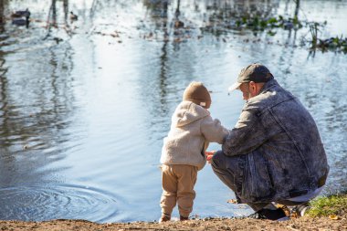 Bir sonbahar parkında sakin bir gölün kenarında duran bir adam ve küçük çocuk. Sarı ve turuncu yapraklı renkli ağaçlar su yüzeyine yansıyor. Aile içi sıcak bir iletişim anı ve sonbahar mevsiminde dışarıda yürüyüş..