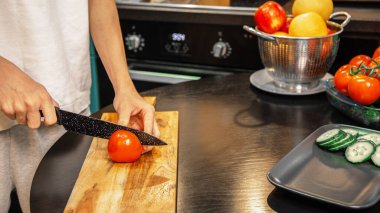 Mutfakta salata hazırlarken tahta kesme tahtasıyla taze domates dilimleyen biri. Taze sebzeler ve meyveler yakınlarda, sağlıklı bir yemek sahnesi oluşturuyor.