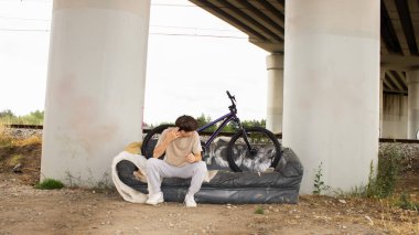 Genç bir çocuk köprünün altındaki eski bir kanepede bisikletle poz veriyor. Endüstriyel bir açık havada gençlik, özgürlük ve yaratıcılık içeren kentsel yaşam sahnesi. 