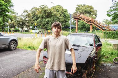 Endüstriyel bir arazide hurdaya dönmüş bir spor arabanın yanında güvenle poz veren genç bir çocuk. Manzara erkekliği, gücü, isyanı ve Z Jenerasyonu sokak tutumunu ifade eder.. 