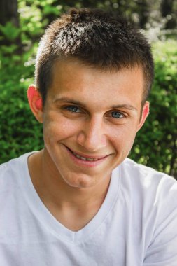 Beyaz tişörtlü, gülümseyen bir gencin yakın plan portresi. Kameraya doğal yeşil arka planda bakıyor. Kısa saçlı ve mavi gözlü mutlu bir genç..